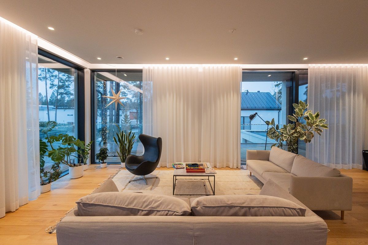 The living room is lit by flush-mounted spotlights and indirectly by LED strips in the ceiling groove that illuminate the day curtains, blocking the view from the outside to the inside during dark hours.